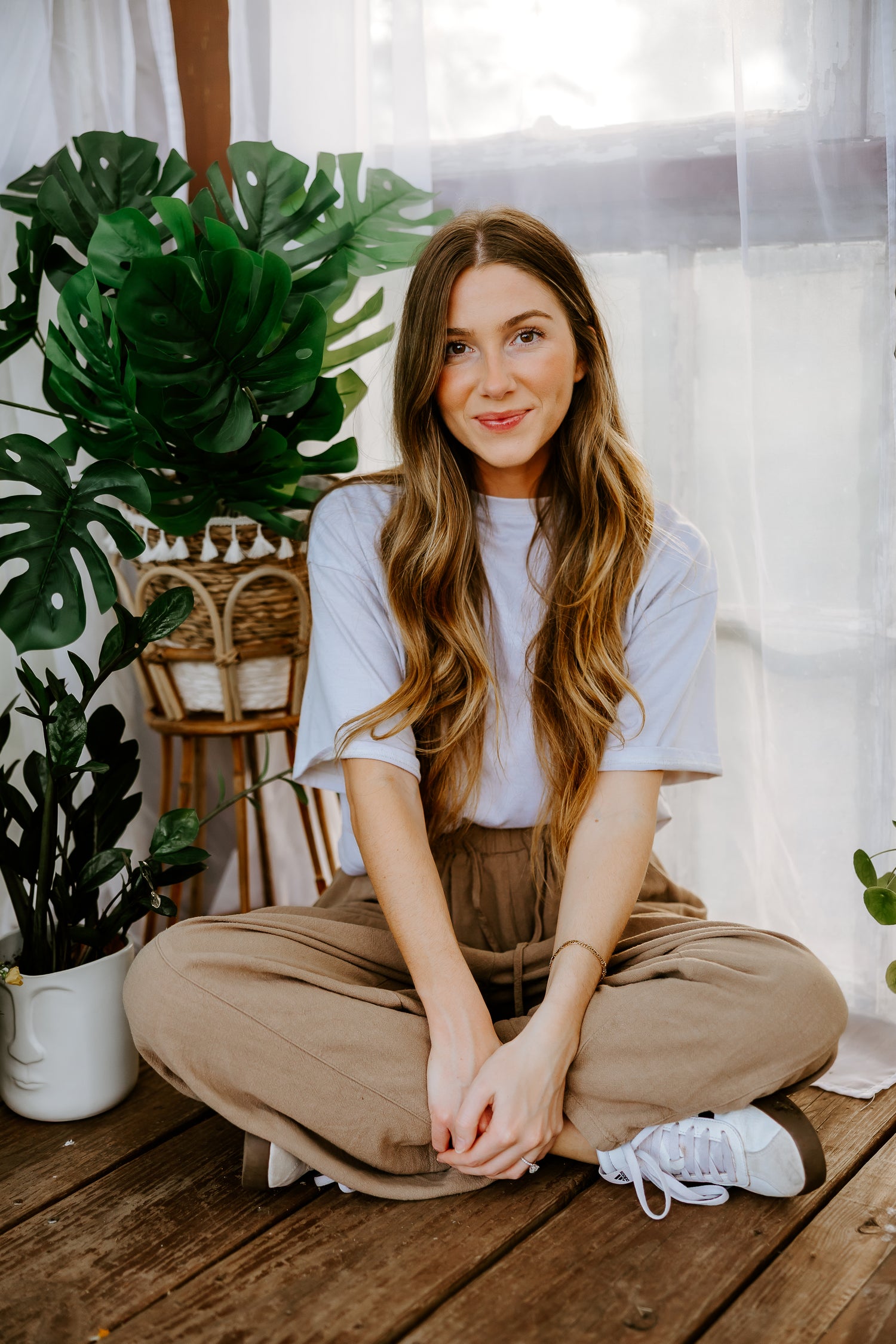 A woman with long wavy brown hair sits cross-legged on a rustic wooden floor in a bright, plant-filled room. She is wearing a light blue short-sleeve t-shirt, brown wide-leg pants, and white sneakers.
