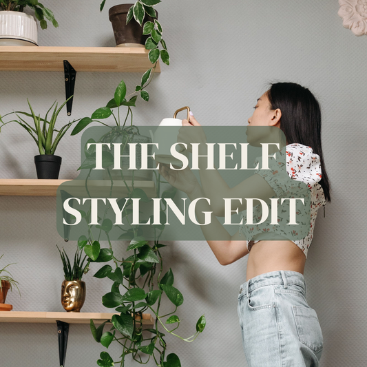 A woman in a floral crop top and light-wash jeans stands beside wooden wall shelves filled with potted plants, holding a white mug. Large overlaid text reads 'THE SHELF STYLING EDIT' across the center.